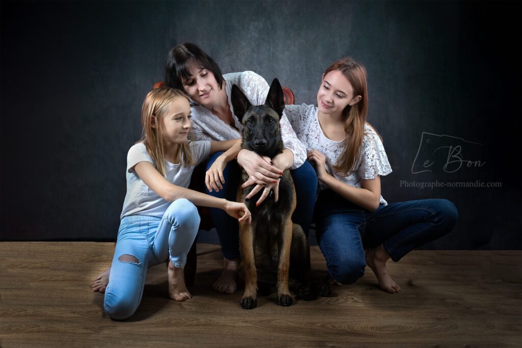 famille d'une maman brune et ses 2 filles assise sur un fauteuil marron en cuir avec un chien malinois  assis à coté