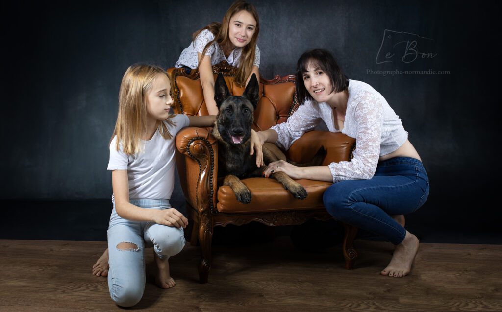 famille d'une maman brune et ses 2 filles assise a coté d'une fauteuil marron en cuir avec un chien malinois allongé dessus